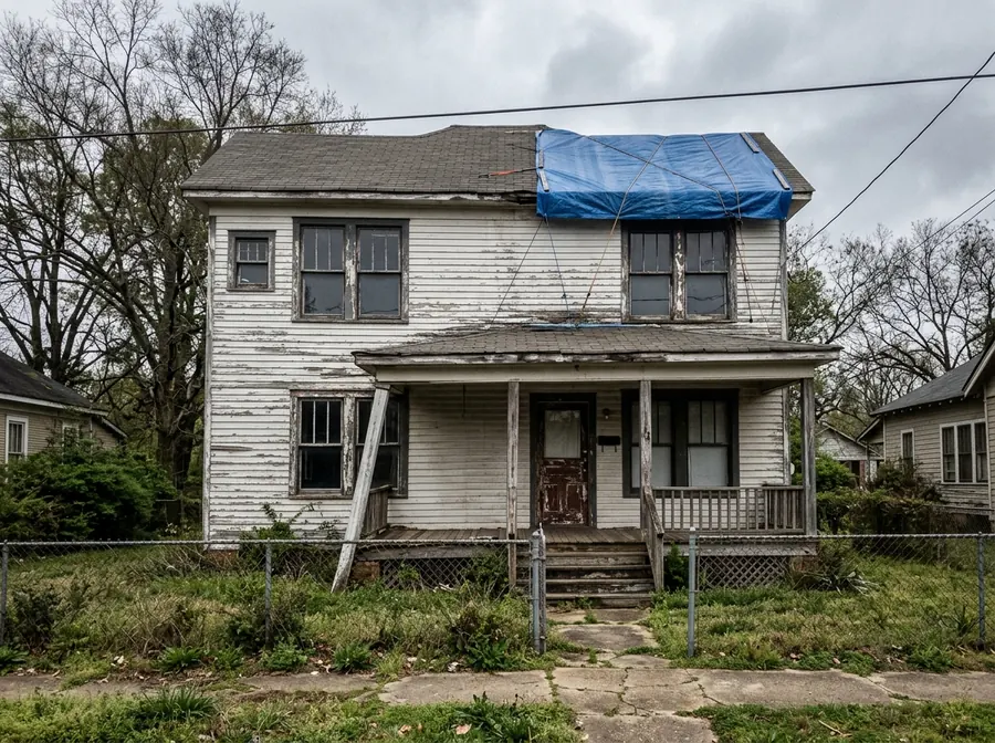 A two storey house with peeling paint, a leaning porch post and a blue tarp over part of the roof.