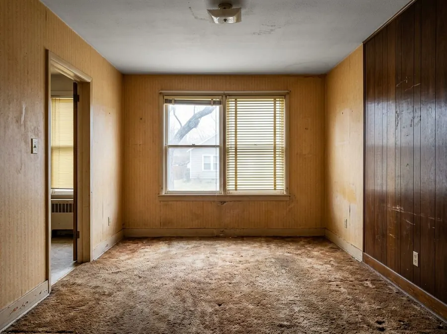 An empty living room with worn brown carpet, yellowed wallpaper and dark wood panelling.